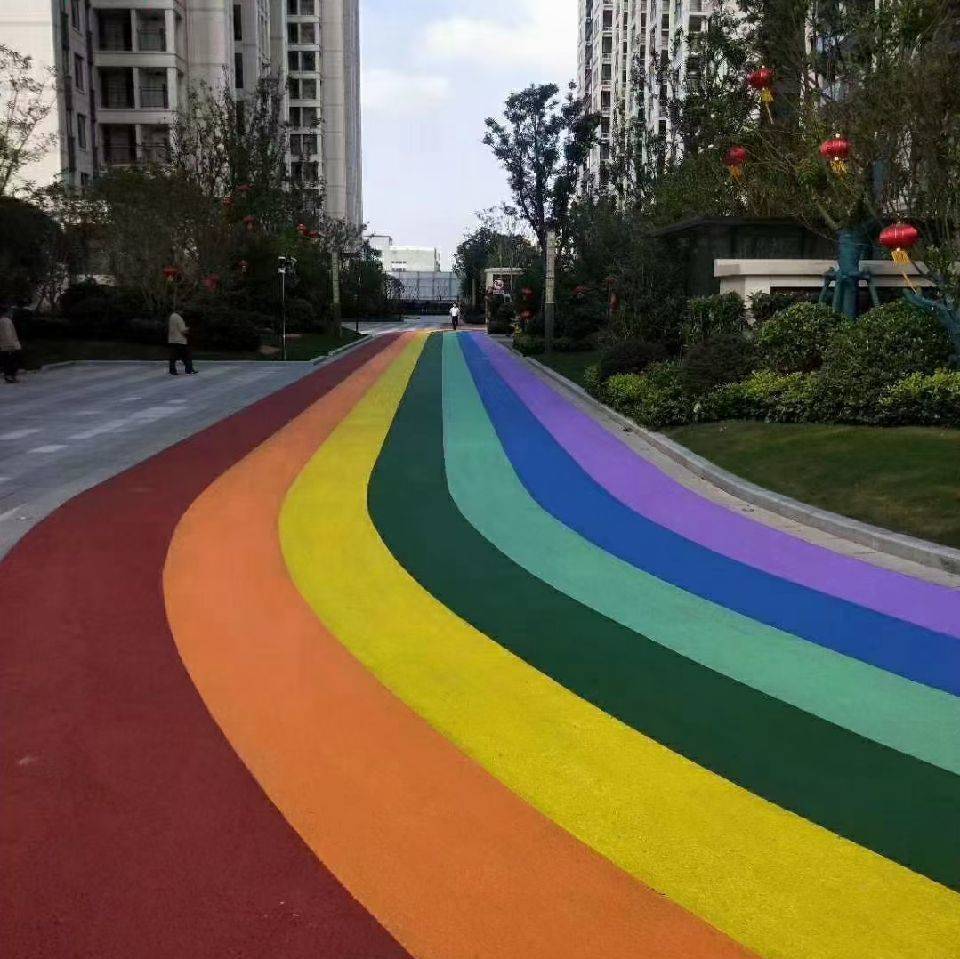 A vibrant rainbow-colored permeable concrete pathway winds through a residential area, featuring distinct red, orange, yellow, green, blue, and purple lanes bordered by greenery and high-rise buildings. A vibrant rainbow-colored permeable concrete pathway winds through a residential area, featuring distinct red, orange, yellow, green, blue, and purple lanes bordered by greenery and high-rise buildings.