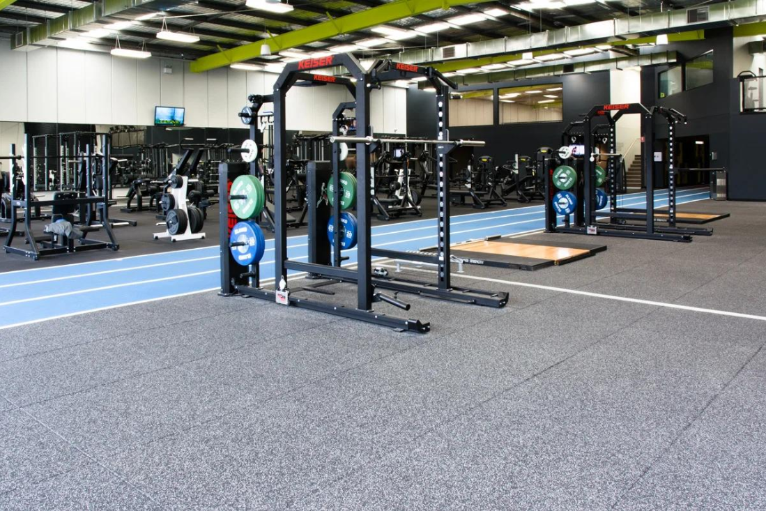 Commercial gym interior featuring gray speckled rubber flooring tiles beneath heavy-duty Keiser power racks and weightlifting platforms, with a blue running track visible in the background.