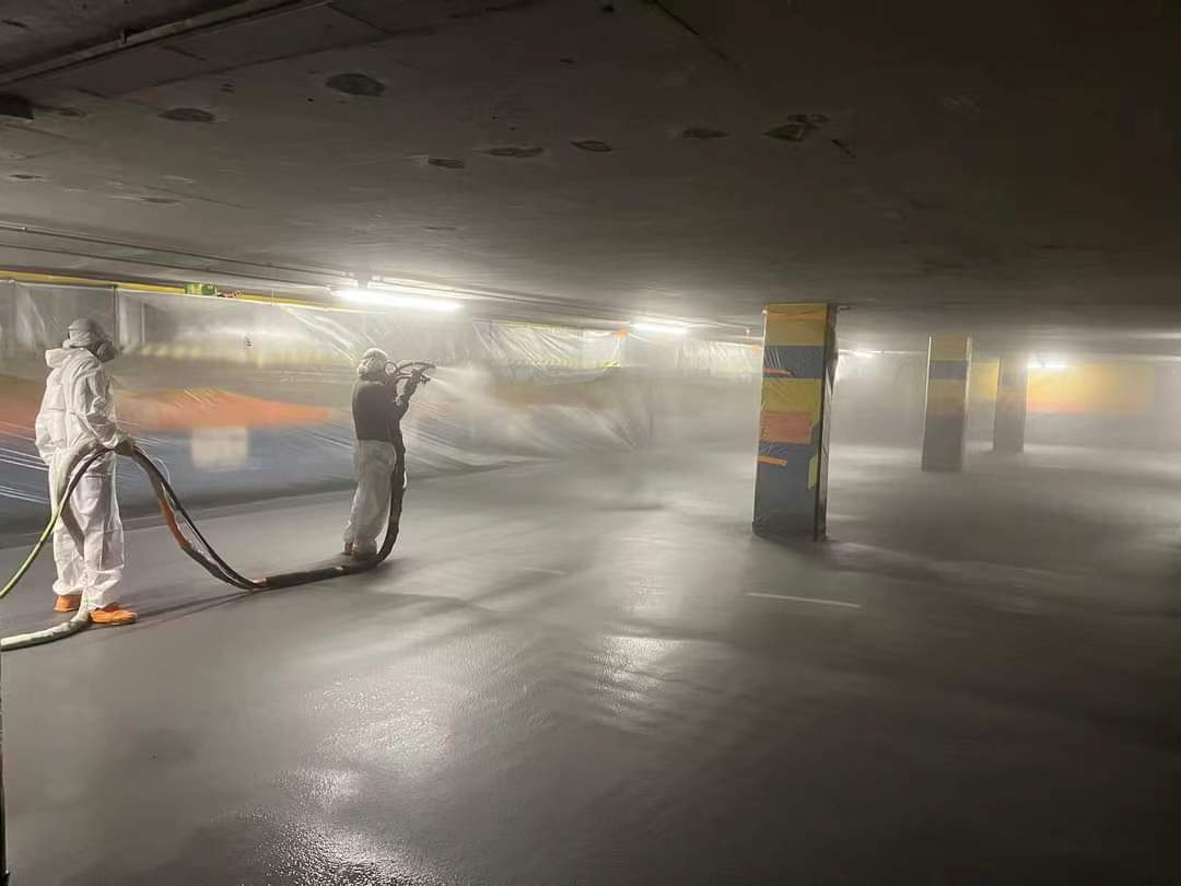 A worker in full protective gear is spraying a polyurea coating onto the floor of an underground parking garage, creating a misty, high-gloss finish that reflects overhead lights, with color-code