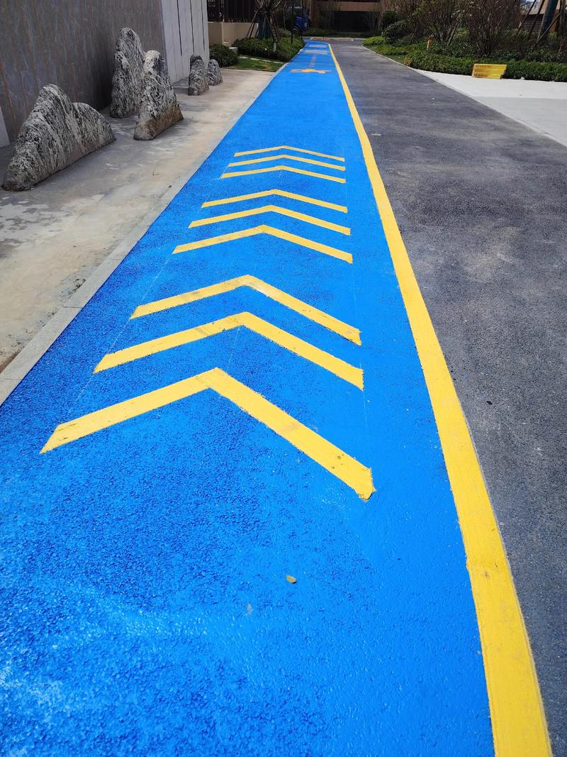 A blue pedestrian or cycling path with yellow directional arrows and boundary lines, running alongside a paved road with decorative rocks and greenery nearby. A blue pedestrian or cycling path with yellow directional arrows and boundary lines, running alongside a paved road with decorative rocks and greenery nearby.