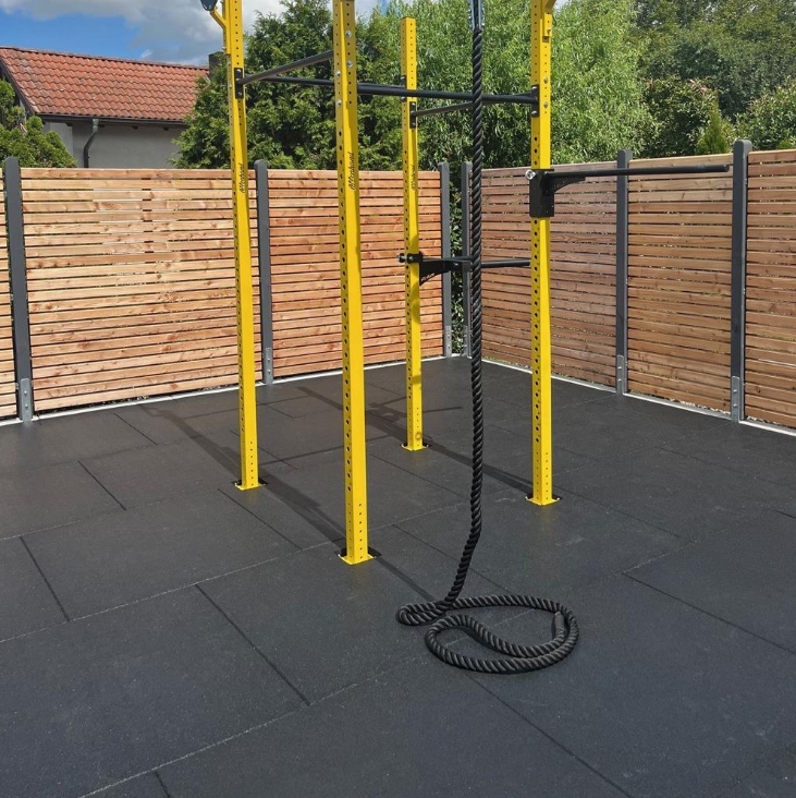 Outdoor functional training zone with black rubber safety tiles under yellow metal rig structures and a climbing rope, enclosed by a wooden fence for private backyard or commercial fitness use.