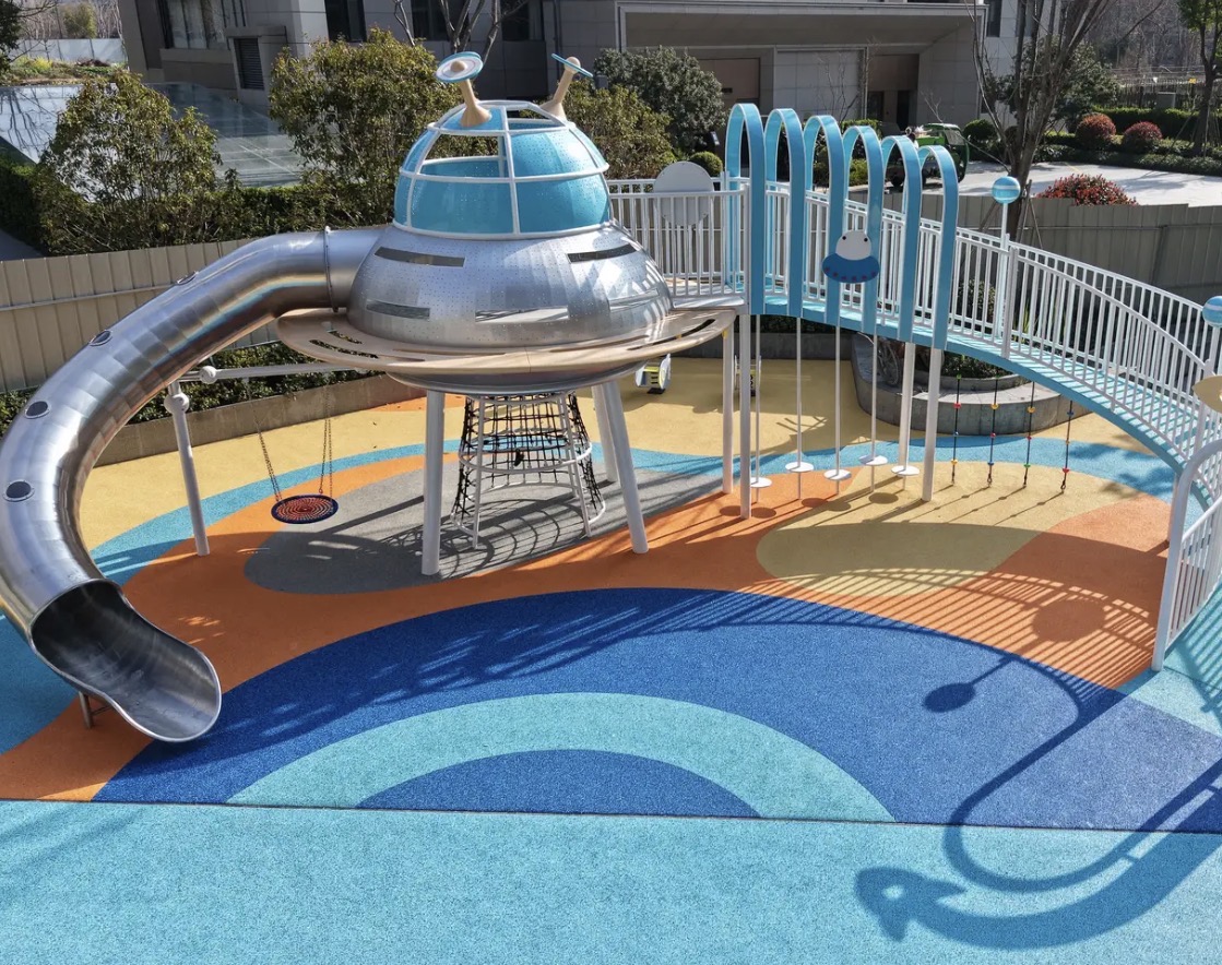 A playground featuring a metallic UFO - shaped slide structure, with the ground covered in colorful EPDM rubber granules in shades of blue, orange, and yellow, providing a safe and vibrant play surf