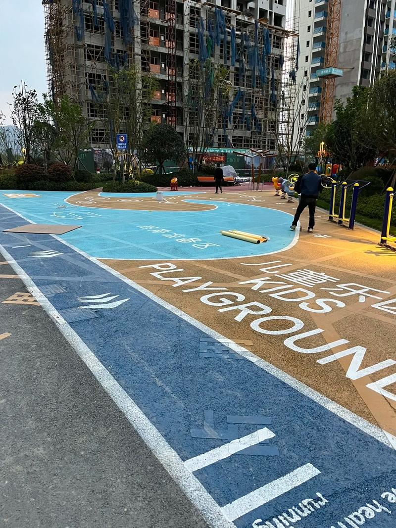 An outdoor children's playground in a residential area, featuring colorful blue and yellow rubber flooring with "KIDS PLAYGROUND" text, surrounded by high-rise apartment buildings under construction and a few people walking nearby. An outdoor children's playground in a residential area, featuring colorful blue and yellow rubber flooring with "KIDS PLAYGROUND" text, surrounded by high-rise apartment buildings under construction and a few people walking nearby.