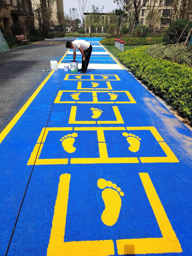 A worker is painting yellow footstep patterns and grid lines on a blue outdoor pathway in a residential area, surrounded by greenery and apartment buildings. A worker is painting yellow footstep patterns and grid lines on a blue outdoor pathway in a residential area, surrounded by greenery and apartment buildings.