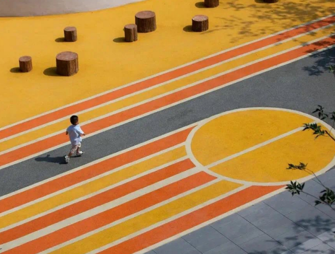 A child walks across a vibrant, multi-colored playground surface featuring bold orange, yellow, and gray stripes with a large circular design, surrounded by tree-stump-style seating elements in a modern outdoor play area. A child walks across a vibrant, multi-colored playground surface featuring bold orange, yellow, and gray stripes with a large circular design, surrounded by tree-stump-style seating elements in a modern outdoor play area.