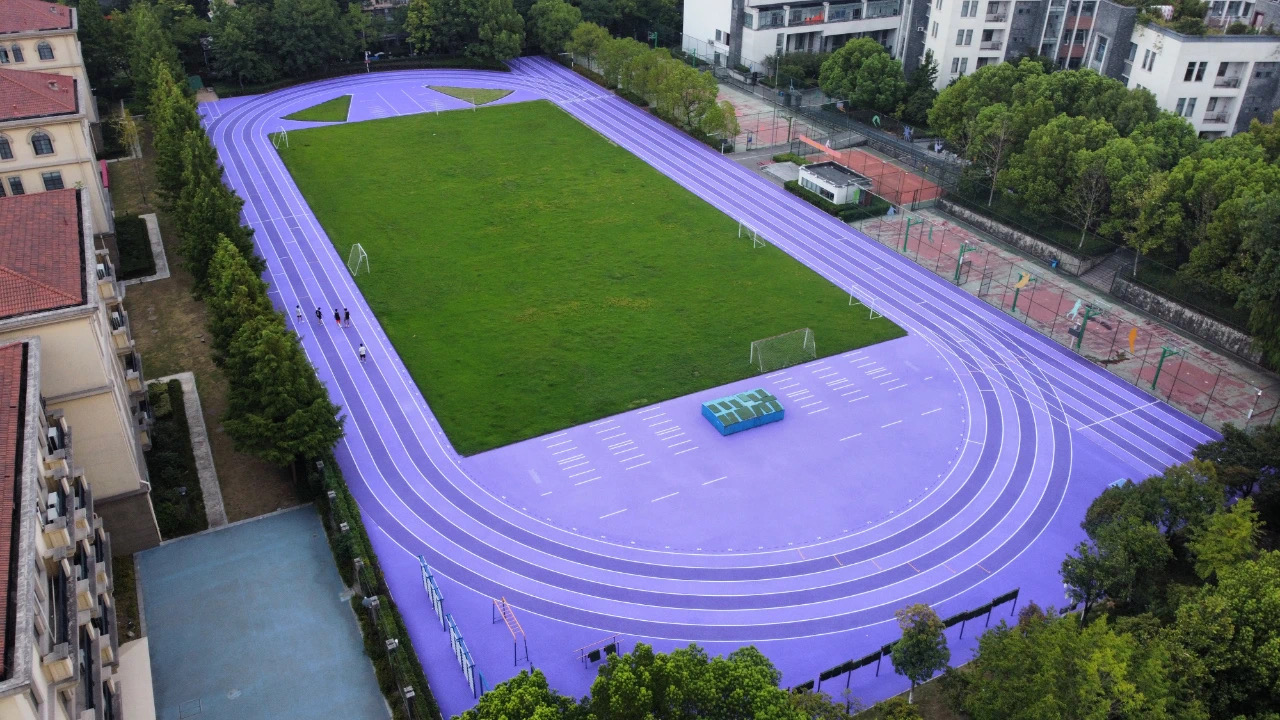 An aerial view of a school athletics track featuring a vibrant purple EPDM granule surface surrounding a green artificial turf field, showcasing a modern and durable sports facility design.