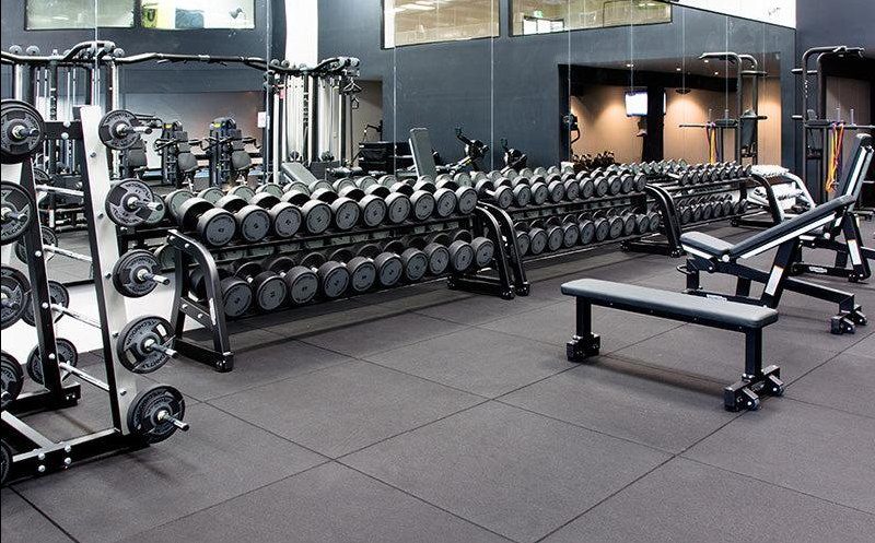 A spacious commercial gym featuring durable black rubber flooring with a speckled texture, designed to withstand heavy weightlifting equipment and high-traffic workouts. Rows of dumbbells and weight benches sit on thick, interlocking gray rubber tiles in a professional gym, highlighting the floorings stability and protective qualities for heavy free-weight areas.
