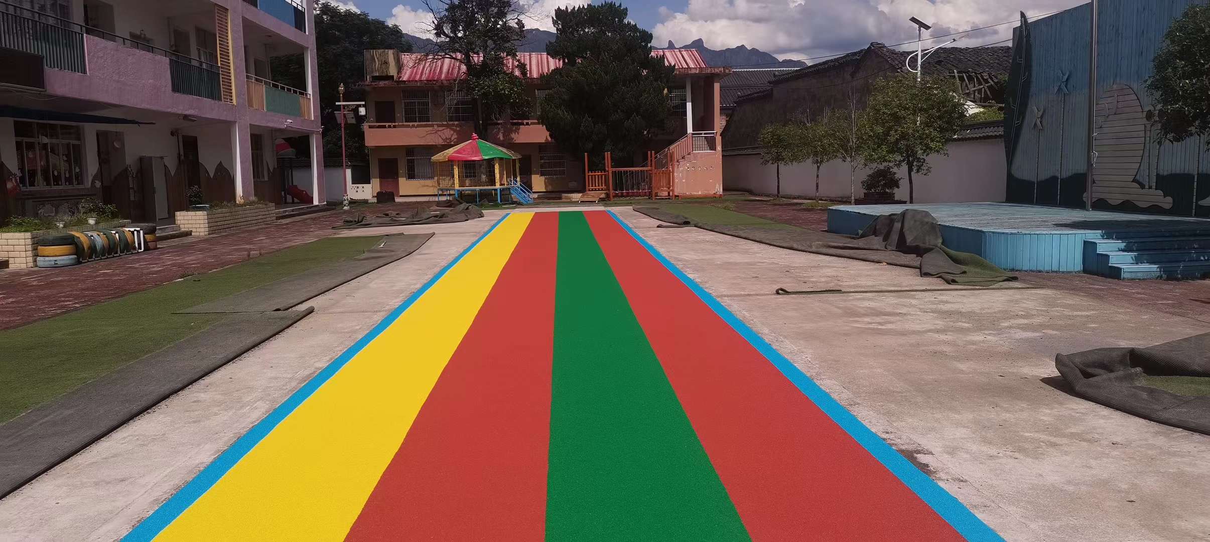 Un vibrante camino de gránulos de EPDM con los colores del arco iris, que presenta franjas llamativas de amarillo, rojo, verde y azul, dispuesto en un área abierta al aire libre rodeada de edificios y vegetación, ideal para parques infantiles.