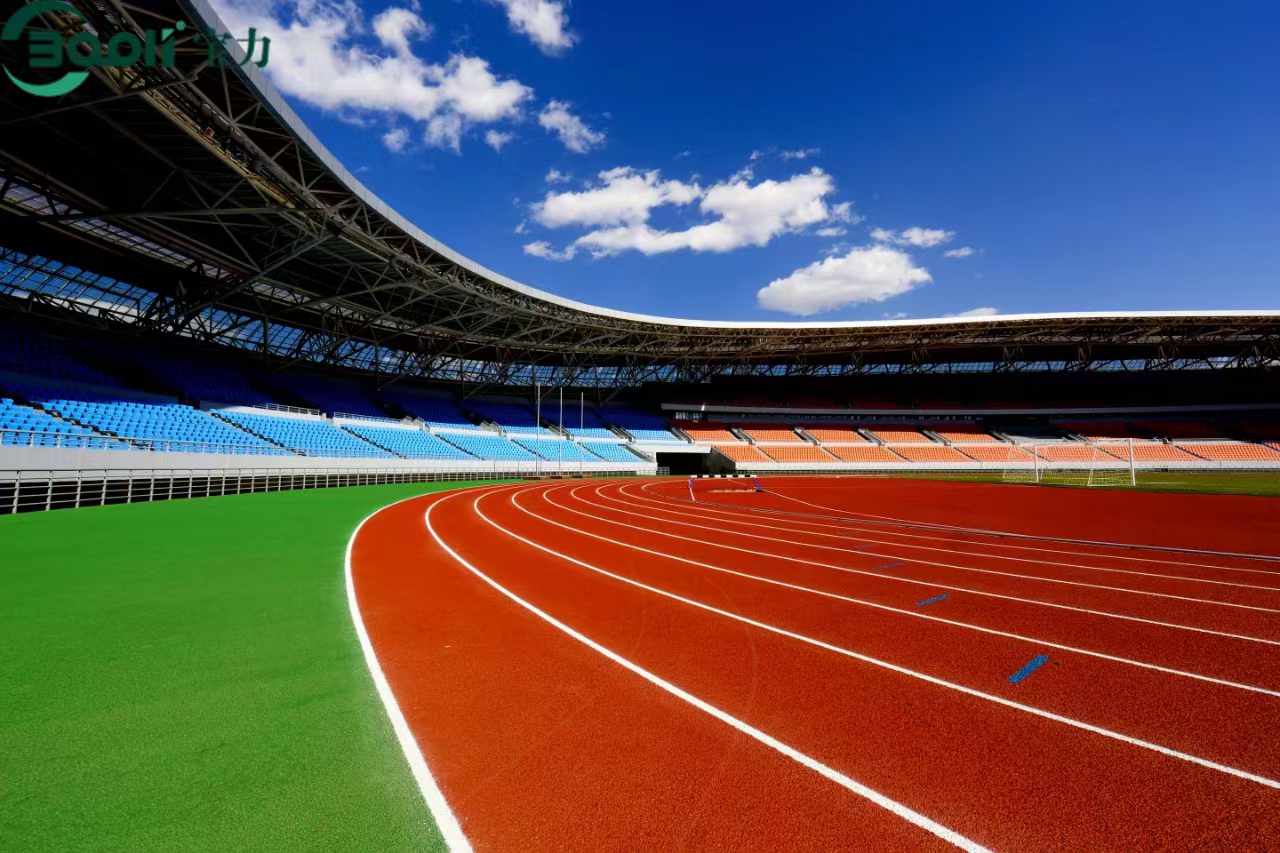 Una vista de gran angular de un estadio profesional con una pista de atletismo sintética de color rojo vibrante con marcas de carril blancas, que se curva a lo largo de un campo de césped artificial verde y exuberante bajo un fondo azul brillante.