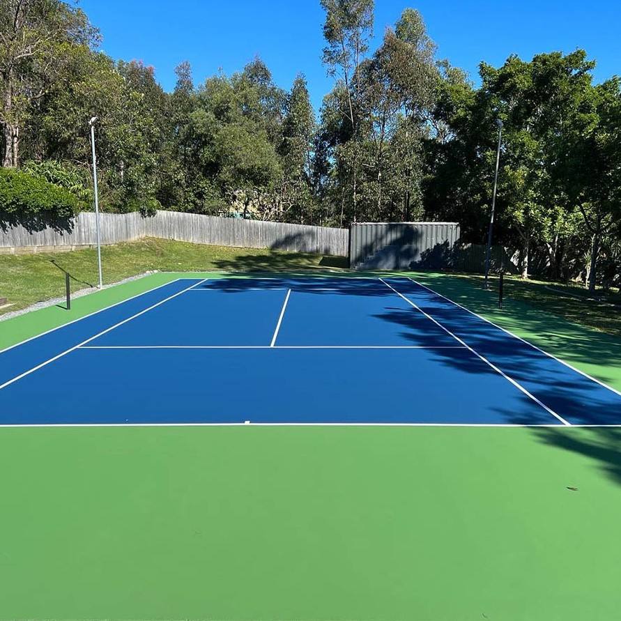 A blue and green tennis court with white boundary lines, surrounded by trees and a fence under a clear sky. A blue and green tennis court with white boundary lines, surrounded by trees and a fence under a clear sky.
