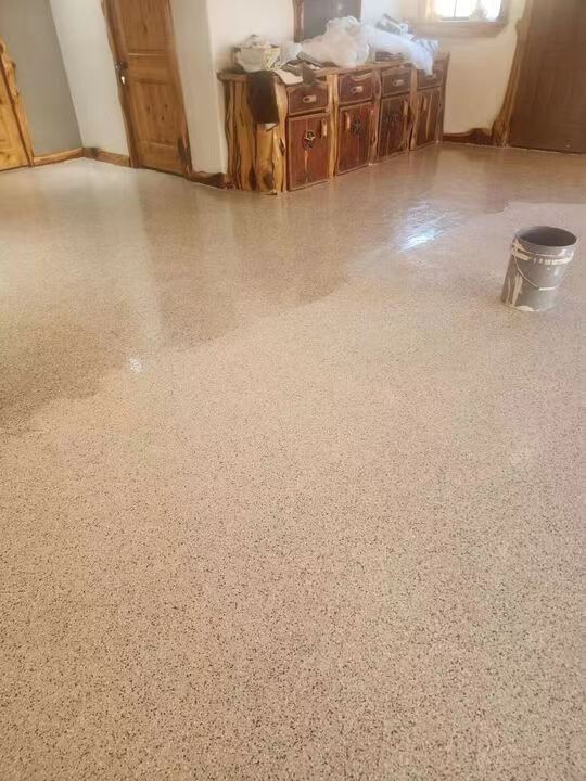 A speckled, seamless polyurea floor coating in a residential interior, with a paint can placed on the surface and wooden furniture in the background, showcasing the materials textured decorative