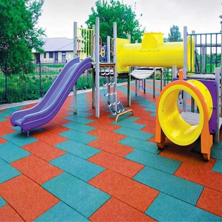 Colorful interlocking rubber safety tiles in red and teal form a shock-absorbing playground surface beneath vibrant play structures including a purple slide and yellow tunnel, designed to protect children during outdoor recreational activities.
