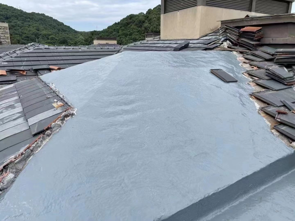 A freshly applied polyurea waterproof coating on a rooftop, forming a seamless, light-gray membrane over the surface, with traditional dark roof tiles visible along the edges and green hills in t