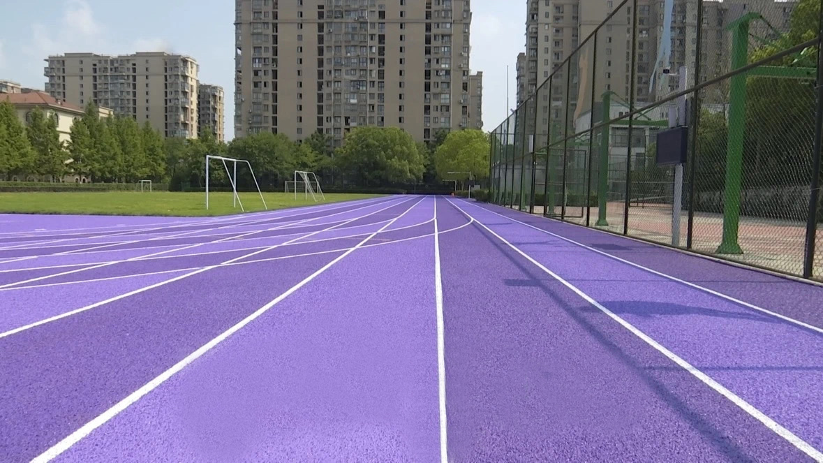A ground-level perspective of a vibrant purple EPDM granule running track with crisp white lane markings, extending toward a green sports field enclosed by fencing and residential buildings in