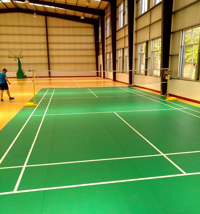 An indoor badminton court with green flooring and white boundary lines, featuring a player in motion near the net under bright artificial lighting. An indoor badminton court with green flooring and white boundary lines, featuring a player in motion near the net under bright artificial lighting.