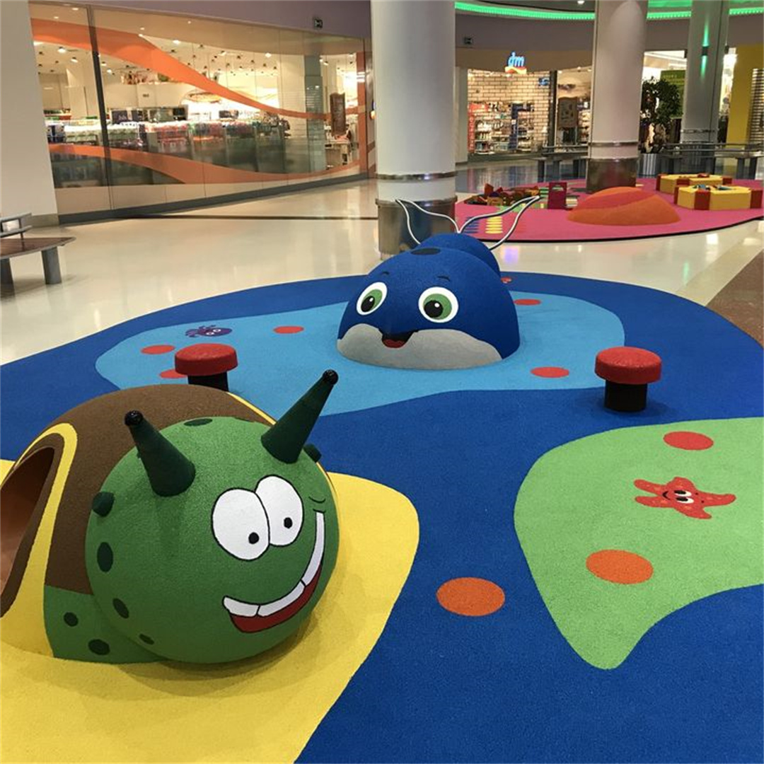 Una vibrante área de juegos interior para niños con coloridos pisos de caucho EPDM en azul, amarillo y verde, con extravagantes estructuras de juego con formas de animales, que incluyen un dinosaurio verde sonriente y