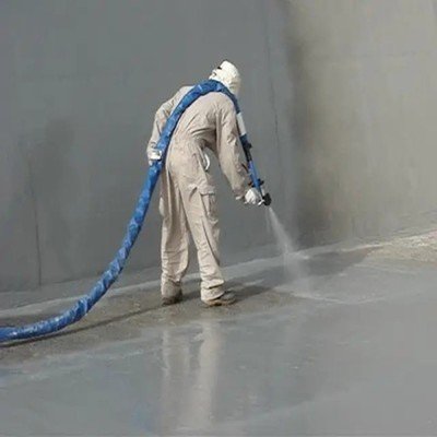 A worker in full protective gear is spray-applying polyurea coating onto a concrete wall and floor junction, using a high-pressure spray gun connected to a blue hose, demonstrating the fast-curin