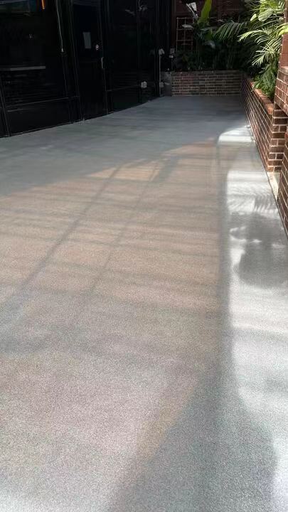 A smooth, light-gray polyurea-coated outdoor walkway with a subtle speckled texture, reflecting sunlight and shadows from nearby structures, demonstrating the material’s durability and aesthetic