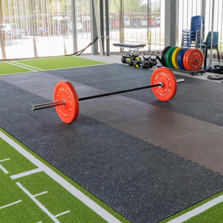 A barbell with bright orange weight plates rests on interlocking black rubber gym tiles, demonstrating the flooring’s shock absorption and durability for heavy lifting zones adjacent to artificial turf.
