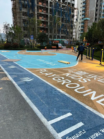An outdoor childrens playground in a residential area, featuring colorful blue and yellow rubber flooring with KIDS PLAYGROUND text, surrounded by high-rise apartment buildings under construction.jpeg