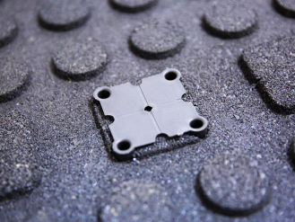 Close-up of a black plastic connector clip securing interlocking rubber gym floor tiles with a textured, dimpled surface for grip and shock absorption..png