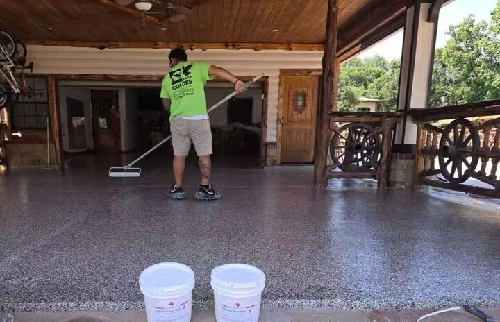 A worker in a green shirt is applying a glossy, speckled polyurea or epoxy coating to a large outdoor patio floor using a long-handled roller, with two white buckets of material in the foreground
