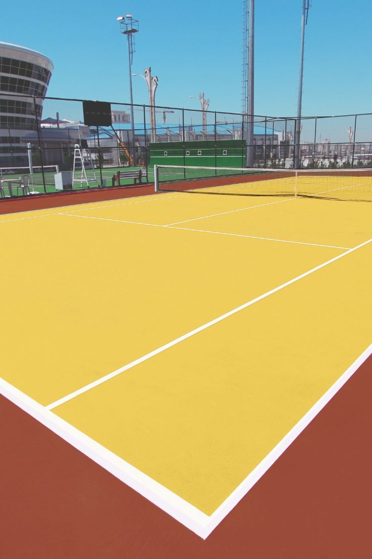 A vibrant yellow and red hard tennis court under clear blue skies, featuring crisp white boundary lines and surrounded by fencing and lighting poles — typical of modern outdoor recreational or