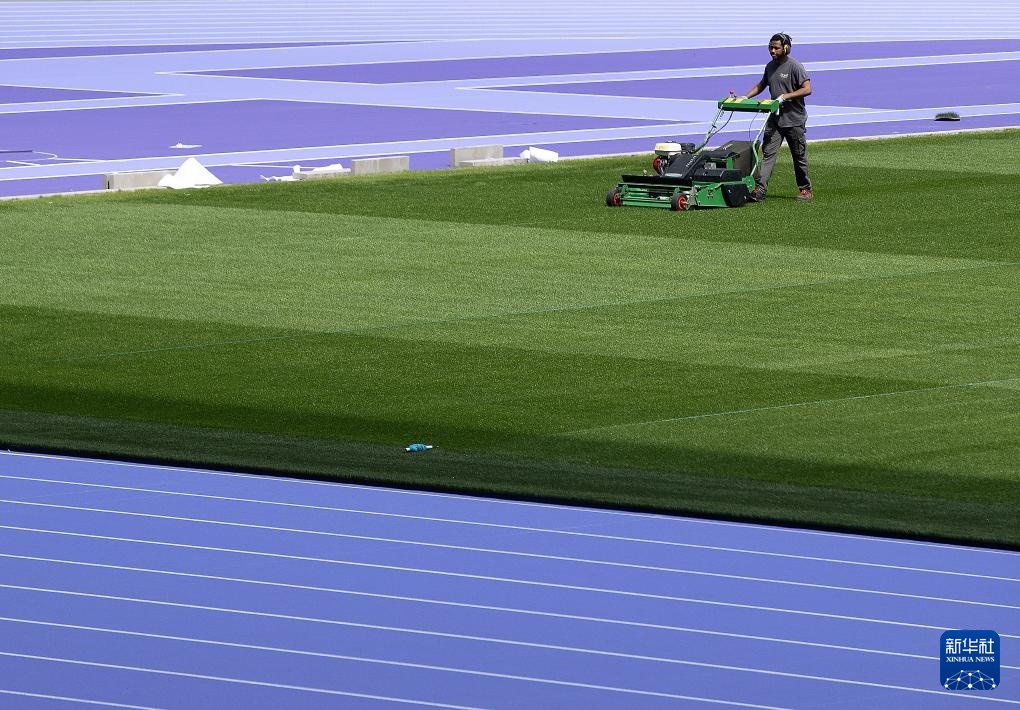 A worker is mowing a lush green lawn with a push mower, bordered by a blue surface with white dashed lines in the foreground and a purple and white patterned area in the background.
