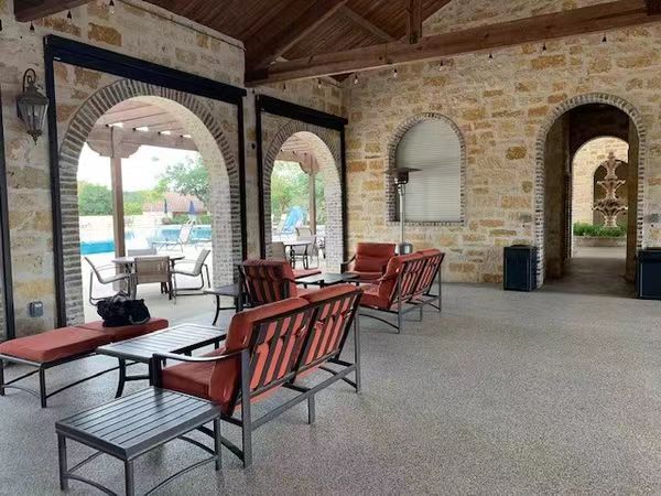 A spacious outdoor patio with a smooth, light-gray polyurea-coated floor, featuring arched stone entryways, red cushioned metal chairs, and a glimpse of a pool in the background, showcasing a dur