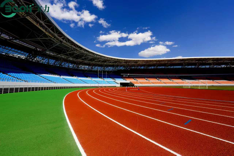 A wide-angle view of a professional stadium featuring a vibrant red synthetic running track with white lane markings, curving alongside a lush green artificial turf field under a bright blue s.jpg