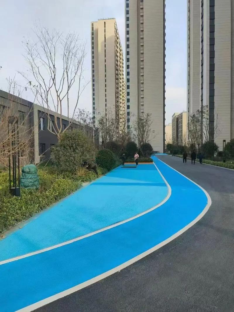 A vibrant blue running track winding through a modern residential complex, flanked by high-rise apartment buildings and landscaped greenery under an overcast sky.