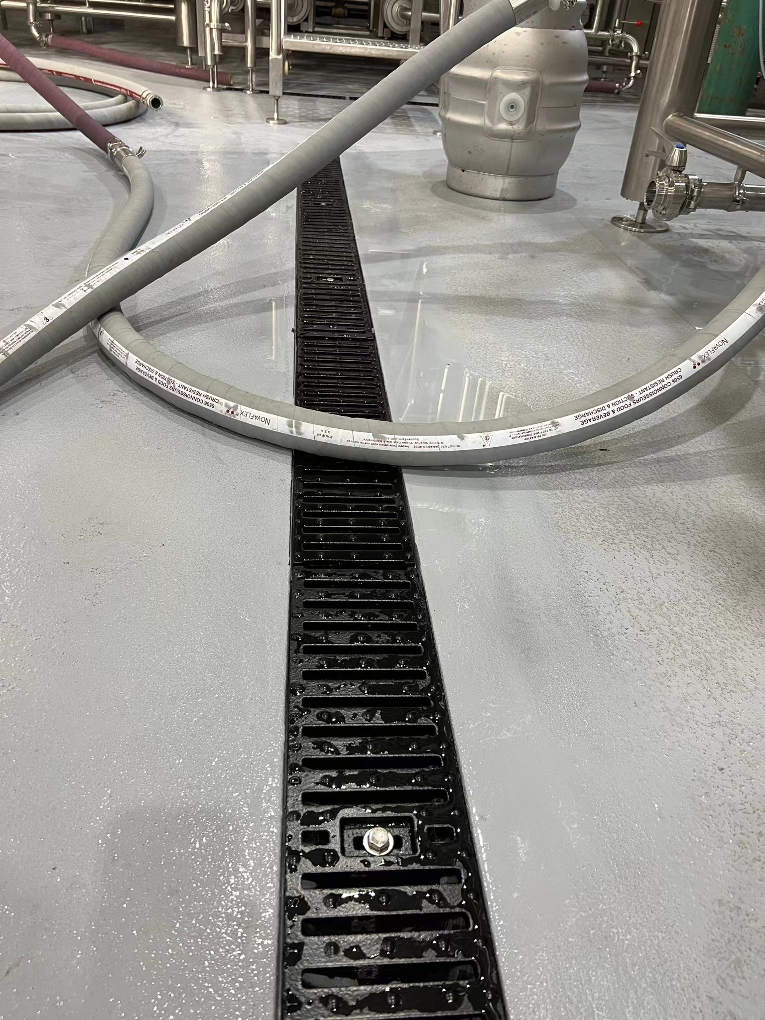 A gray polyurea-coated industrial floor featuring a central black linear drainage grate, with flexible hoses and stainless steel equipment visible in the background, highlighting a hygienic and c